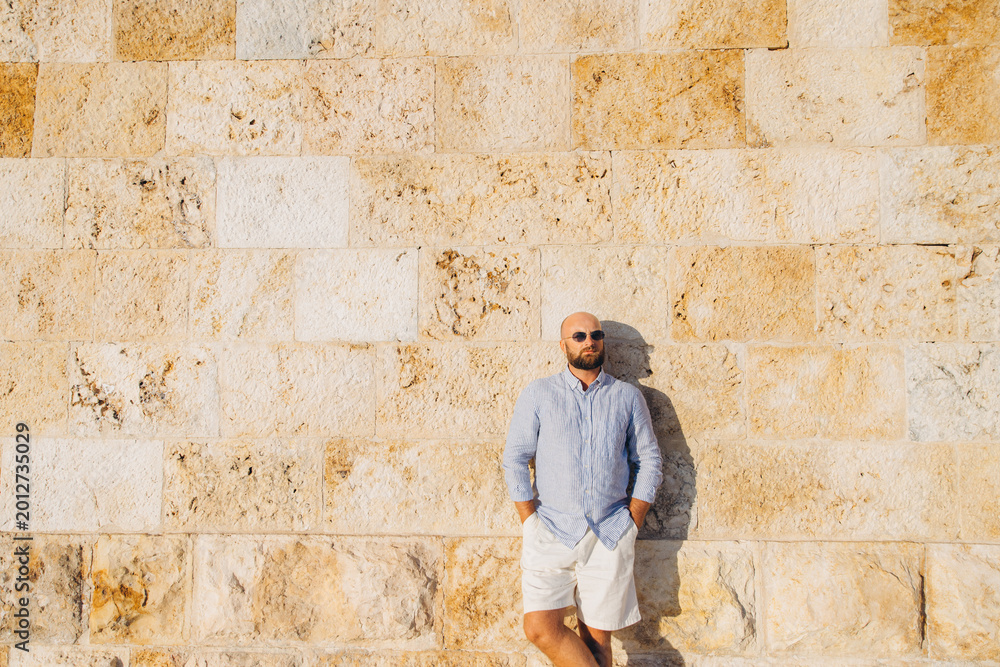 Obraz premium Bald man wearing sunglasses and casual summer clothes standing relaxed against a textured stone wall under warm sunlight