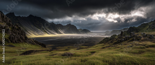 Dramatic coastal mountains with black sand and green grass under moody clouds; cinematic landscape style, wild nature concept, ideal for travel, tourism, posters, backgrounds and editorial visuals