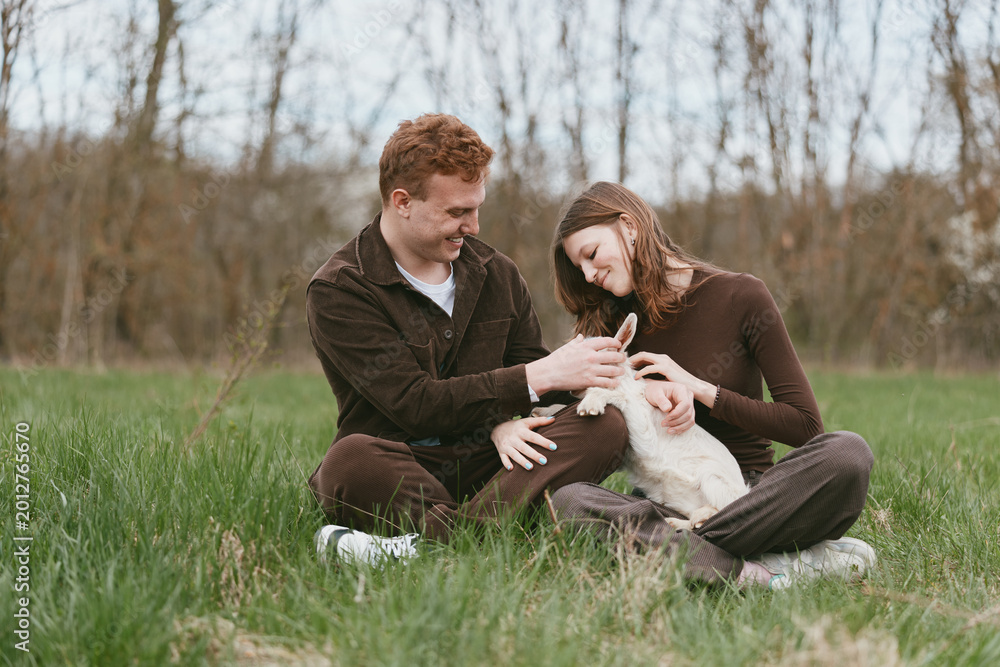 Obraz premium A smiling couple sits on the grass in a park, cradling a young goat kid between them. A candid lifestyle moment highlighting connection, nature, and playful companionship.