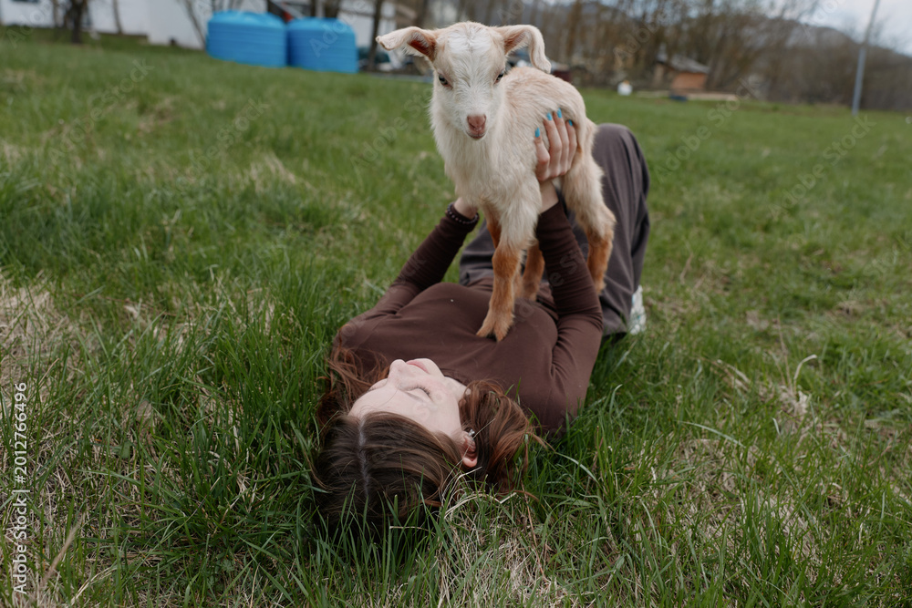 Naklejka premium Young woman lies on grass holding a small goat above her with a joyful expression in a natural outdoor setting during a casual lifestyle moment.