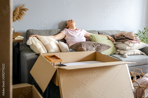 Obraz na płótnie Tired middle aged woman resting on sofa among cardboard boxes and packed belongings after relocation