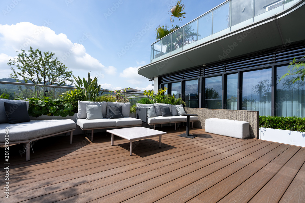 Obraz premium Modern rooftop terrace with wooden deck, outdoor sofas, green plants and glass railing under blue sky