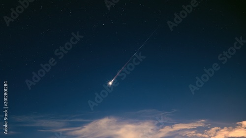 Bright shooting star streaking across the dark night sky with a glowing trail above soft clouds in a long exposure astronomical photograph capturing a celestial event