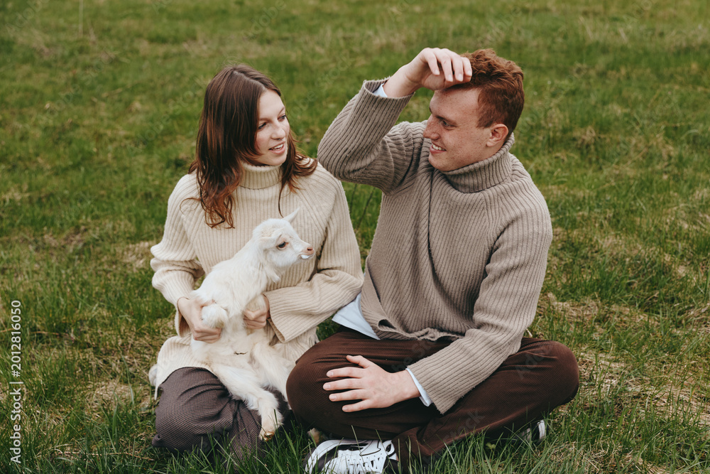 Fototapeta premium Couple sitting on grass with a white goat kid, wearing sweaters, enjoying a relaxed outdoor lifestyle moment with nature, affection, and gentle companionship in warm lighting, cozy ambiance.