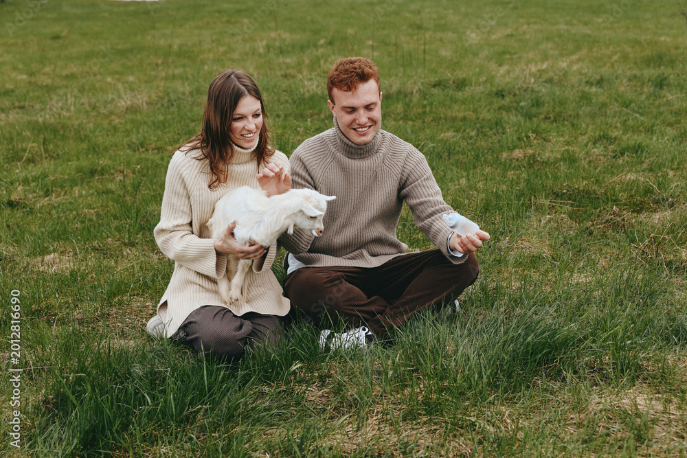 Naklejka premium Couple sitting on grass with a playful goat kid, sharing a gentle moment outdoors. Cozy sweaters, natural light, warm countryside mood. They interact tenderly, suggesting lifestyle, wellness, and