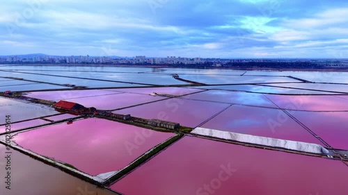High angle shot of rectangular salt pans filled with pink brine under a cloudy sky near a coastal city with white salt heaps. Aerial view of pink salt evaporation ponds in Burgas Bulgaria.