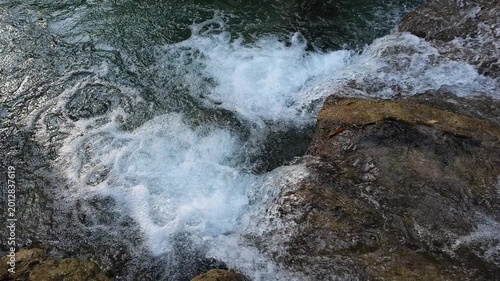 Cinematic slow motion of crystal clear water flowing over rocks in a natural stream. Bubbling white foam and emerald ripples splashing in a tropical forest waterfall. High quality 4K nature background
