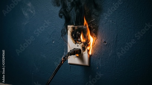 Smoldering electrical outlet catches fire with bright orange flames and thick black smoke emerging against a dark moody blue textured wall.