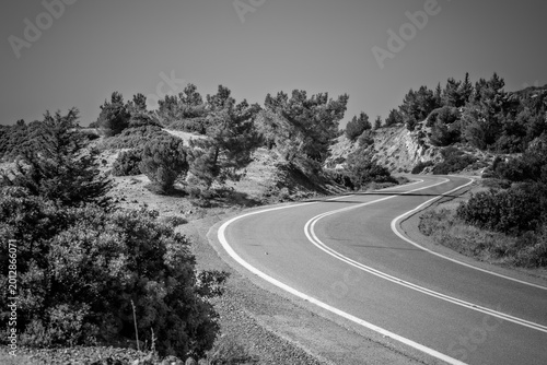 Fotografie Rhodes, Greece: Paved two-lane highway with distinct white dividing lines curving through a hilly arid landscape populated with scattered pine trees and low scrubland brush