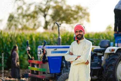 Fotografi young indian farmer standing with tractor at agricultural field