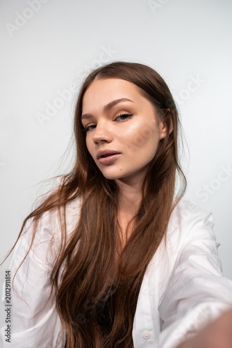 Young woman with long brown hair posing confidently against a white background, wearing a white shirt