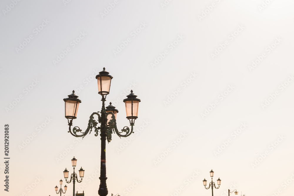 Obraz premium Ornate Lamppost With Distant Lights and Open Sky. A decorative Venice lamppost is isolated against a soft bright sky.