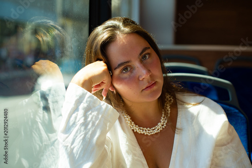 Bild auf Leinwand A young woman wearing a white shirt and pearl necklace sits inside a bus leaning her head on her hand near the window
