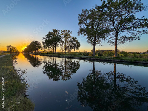 Fotografi Peaceful canal at sunset with perfect reflections in Rijkevorsel, België - Golde