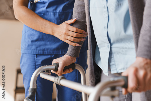 Obraz na plátne Close up of senior woman walking with walker while nurse helps her at home, care