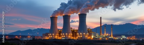 Industrial power plant emitting smoke against a sunset with mountains in the background.