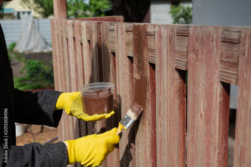 Person painting wooden fence with brush and container in hand on sunny day