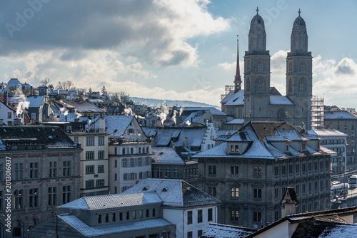 The twin towers of Grossmunster cathedral rising above snow-covered rooftops with smoking chimneys in Zurich