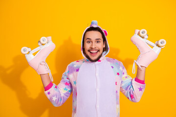 Happy guy in pastel unicorn pajama jumpsuit holds pink roller skates against a bright yellow background for a fun playful home lifestyle photo