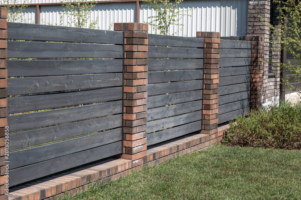 Obraz premium Dark horizontal wooden fence planks with red brick pillars and base near green lawn, residential property boundary and perimeter wall