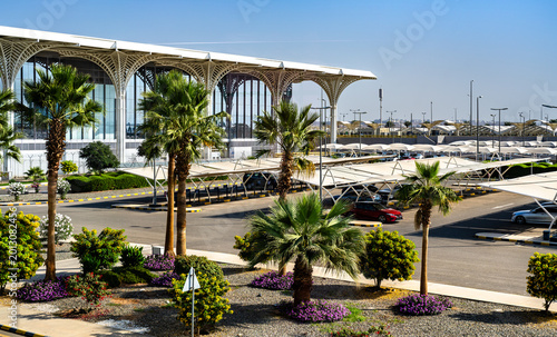 The modern exterior design and landscaping of Prince Mohammad bin Abdulaziz International Airport in Madinah, Saudi Arabia, showing the terminal and parking area.