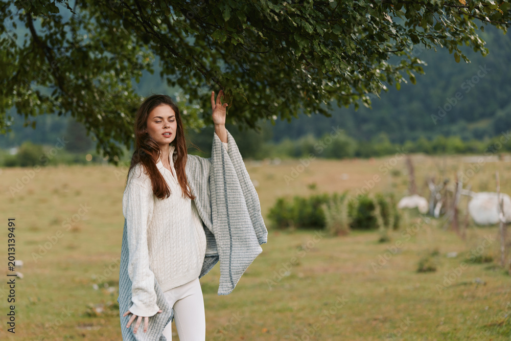 Naklejka premium A woman in a cozy knit sweater stands beneath a leafy tree in an open field, holding a soft blanket over her arm. Bright natural light highlights a calm, casual lifestyle moment outdoors.