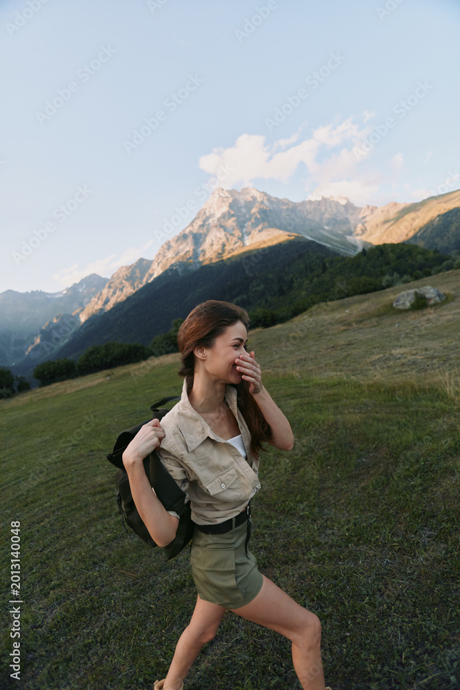 Fototapeta premium Young woman with backpack hikes across a grassy mountainside, enjoying a sunny day and fresh air, in casual utility outfit as mountains loom in the background.