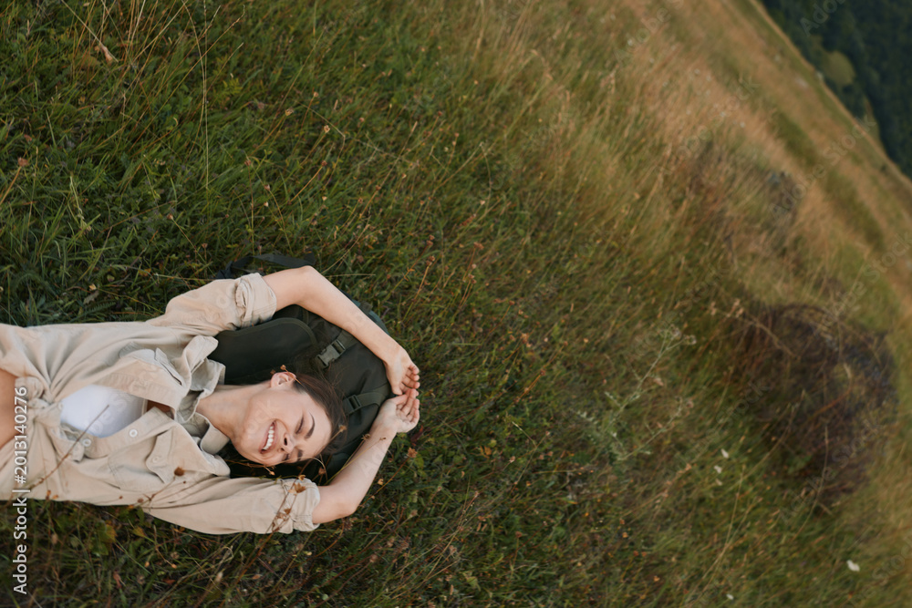 Naklejka premium Young woman rests on grass in a sunlit meadow, eyes closed, wearing a light beige shirt, conveying calm relaxation and connection with nature.