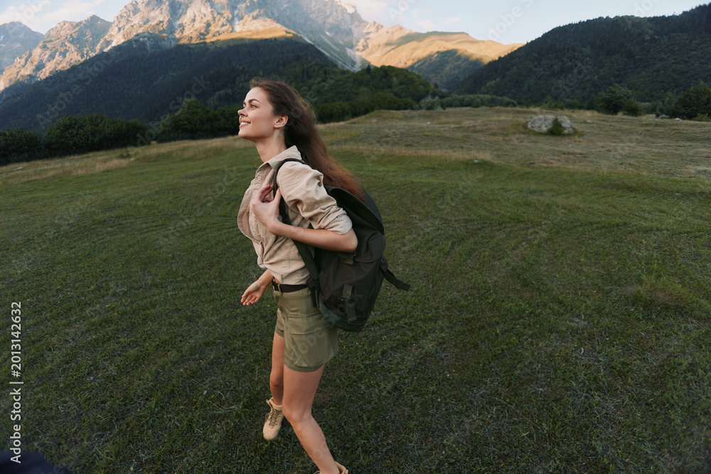Naklejka premium A woman hikes outdoors with a backpack through a grassy valley, with mountains in the distance, conveying adventure, exploration, and a sense of freedom.