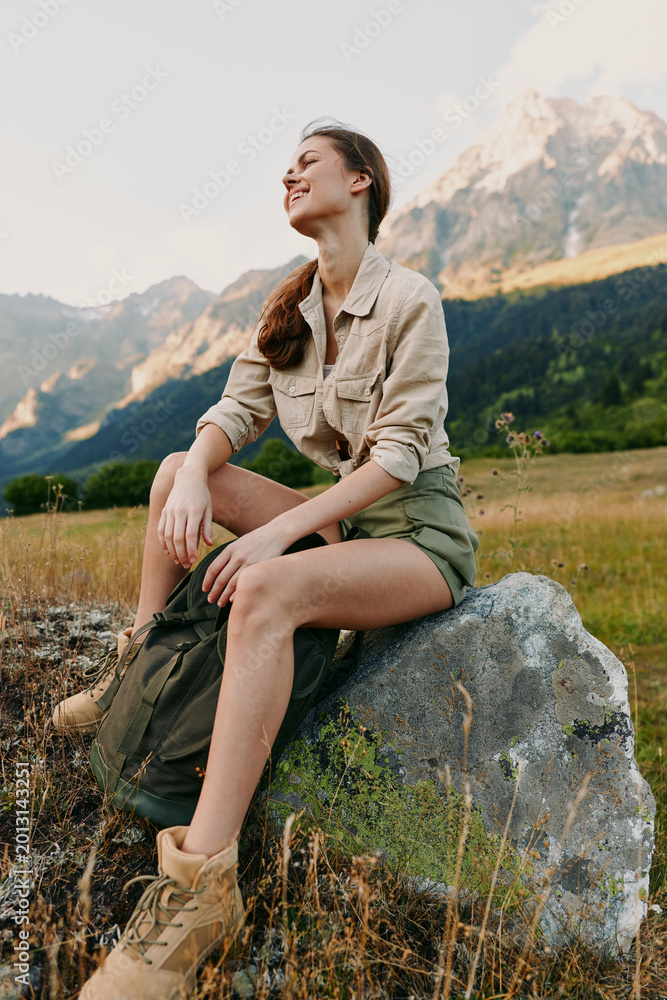 Fototapeta premium Young woman sits on a rock in a mountain meadow, wearing a beige shirt and green shorts, enjoying the fresh air, distant peaks, and a relaxed, carefree mood.