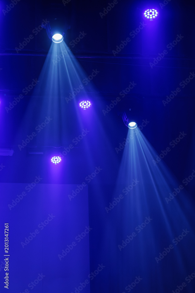Fototapeta premium Blue and purple stage lights cast bright beams through haze from a dark ceiling rig, creating a dramatic performance background with copy space