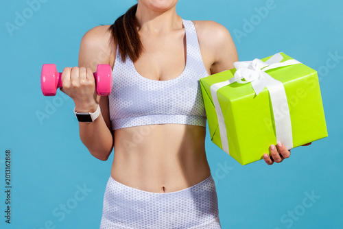 Faceless woman holding pink dumbbell and green gift box to represent gym membership gift isolated over blue background