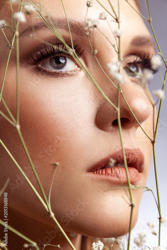 Extreme close up of woman face with professional makeup and long lashes looking through dried flowers isolated over gray background