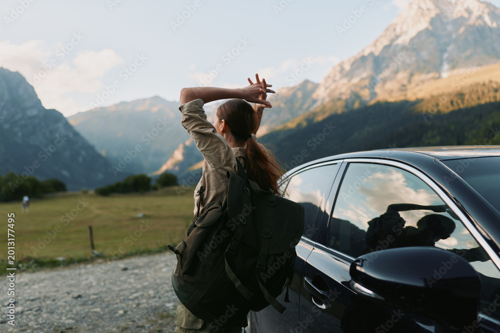 Fototapeta premium Young woman with a backpack stands beside a parked car in a valley, hands behind her head, gazing at distant mountains, enjoying a tranquil outdoor moment of freedom
