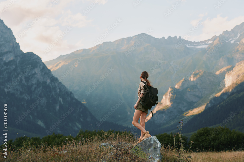Naklejka premium Hiking woman with backpack stands on a rock in a mountain landscape, enjoying nature and solitude, outdoor adventure mood, bright sky. warm sunlight, distant peaks, peaceful