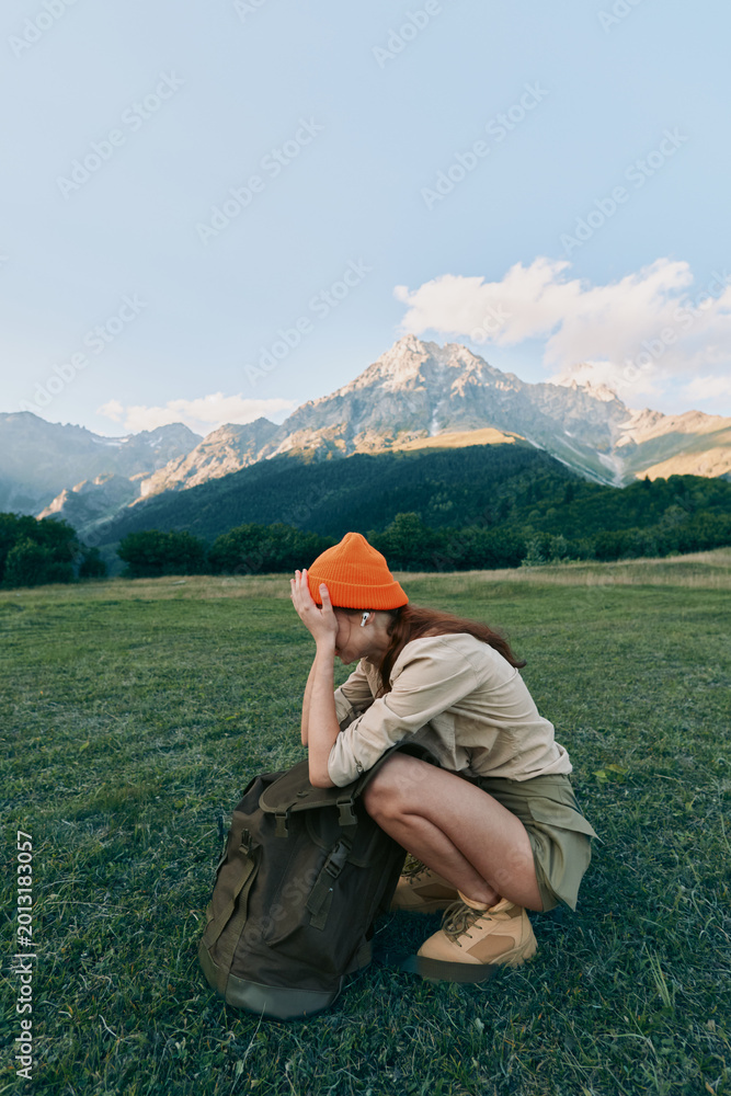 Naklejka premium Young woman crouches in a sunlit meadow with a backpack, shielding her eyes as rugged mountains rise in the background.
