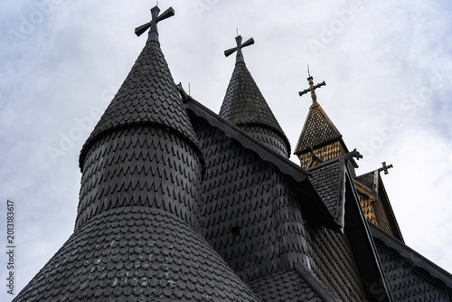 Traditional Norwegian stave church surrounded by spring countryside and forested hills under dramatic cloudy sky in Telemark, Norway. Dark wooden medieval-style architecture. 