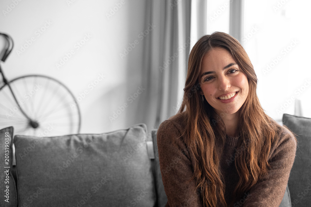 Naklejka premium Smiling young woman sitting on a sofa in a cozy living room, wearing a casual sweater and looking relaxed and approachable; natural light and warm tones create a comfortable, friendly atmosphere.