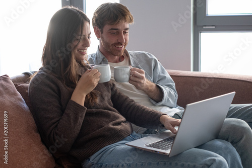 Fotografie A relaxed couple sits closely on a sofa, sharing coffee while browsing on a laptop