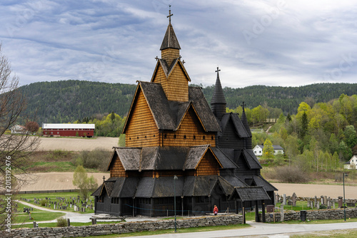 Traditional Norwegian stave church surrounded by spring countryside and forested hills under dramatic cloudy sky in Telemark, Norway. Dark wooden medieval-style architecture.