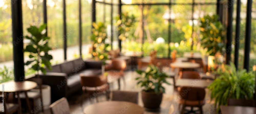 Fototapeta premium A cozy cafe interior featuring blurred tables and chairs surrounded by lush green plants. Large windows allow natural light to fill the space.
