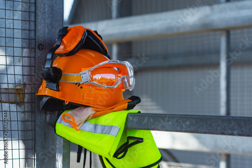 Fotografi Orange hard hat with attached earmuffs and safety goggles on a yellow high visibility safety vest hanging on metal railing at construction site