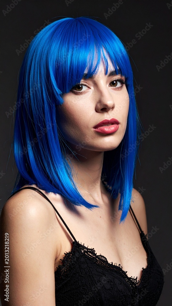 Fototapeta premium A beautiful young woman with bright blue hair wearing a stylish black lace dress looking at the camera against a dark minimalist background.