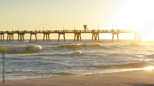 Картината върху платно Sunset over beach with people walking, pier in background, creating a serene coa