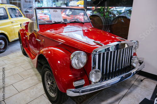 Close-up of shiny red vintage convertible car parked indoors on tiled floor. Chrome grille, round headlights, and wire-spoke wheels are prominent. Yellow vintage car and wooden barrels are visible in