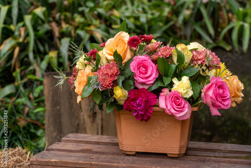 Beautiful colorful flower arrangement with pink, orange and yellow roses in a terracotta pot on a rustic wooden bench with garden background.