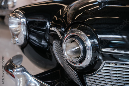 Detailed low-angle view highlights front right fender, headlight, and grille of vintage black automobile. Glossy paint reflects light, showcasing chrome accents and classic design elements.