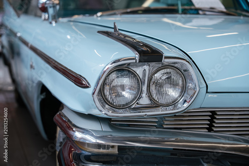 Detailed frontal view of classic pale blue automobile, showcasing dual round headlights, chrome grille, and shiny chrome bumper. Decorative chrome trim and hood ornament enhance vintage appeal.