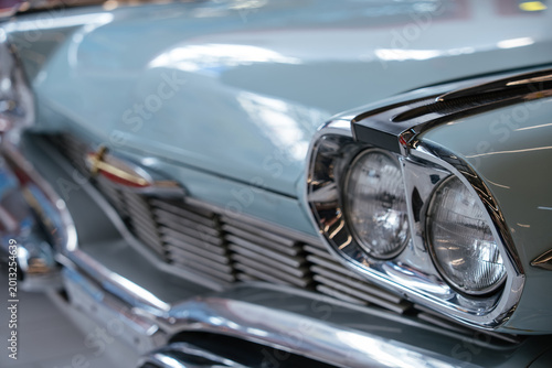 Detailed close-up view of front section of light blue vintage automobile. Polished chrome headlights and grille reflect surroundings, emphasizing classic design in a softly blurred indoor setting.