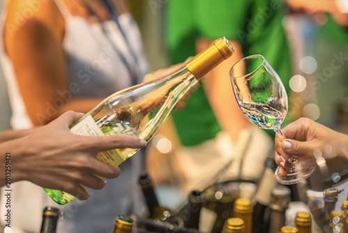 White wine tasting at indoor event with bottle and glass close-up. Wine being poured from glass bottle into tasting glass, blurred people, bottles, warm lights, shallow depth of field in background